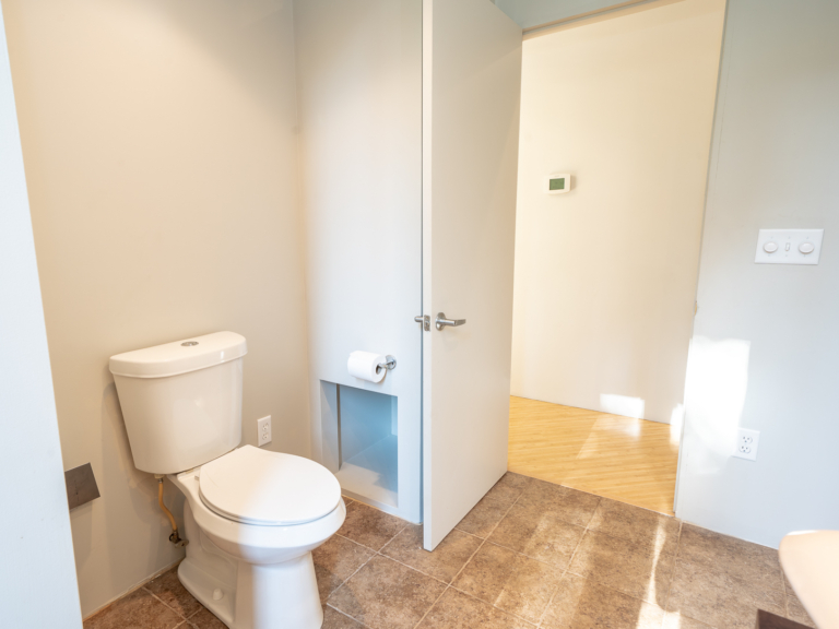 Toilet and storage area of secondary bathroom in modern renovation home project by The RBT Group featuring tiled flooring, clean color palette, and dimmed lighting panel.