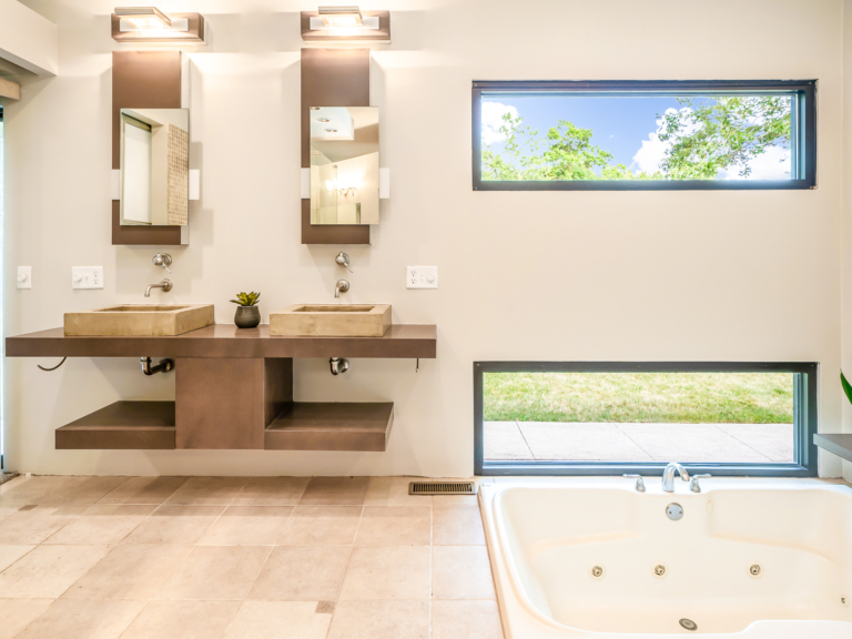 Double concrete sinks, unique mirrors and lighting, a in floor soaking tub, and horizontal feature windows of primary bathroom in modern renovation home project by The RBT Group.