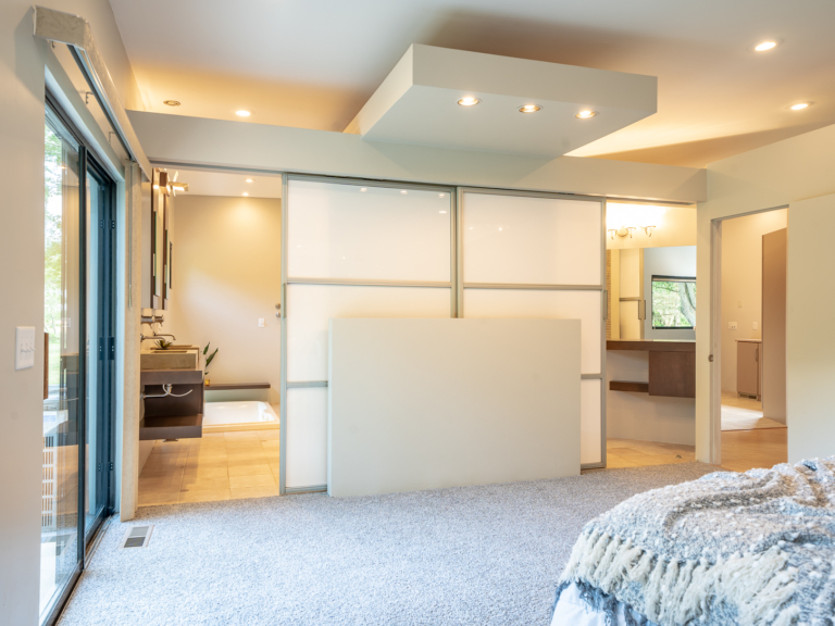 Feature wall in primary bedroom leading into bathroom in modern renovation home project by The RBT Group, with sliding frosted glass doors and wall space for decor.